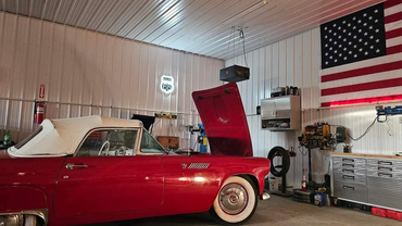 Red classic car in a well-lit garage, hood open. American flag and tools visible.