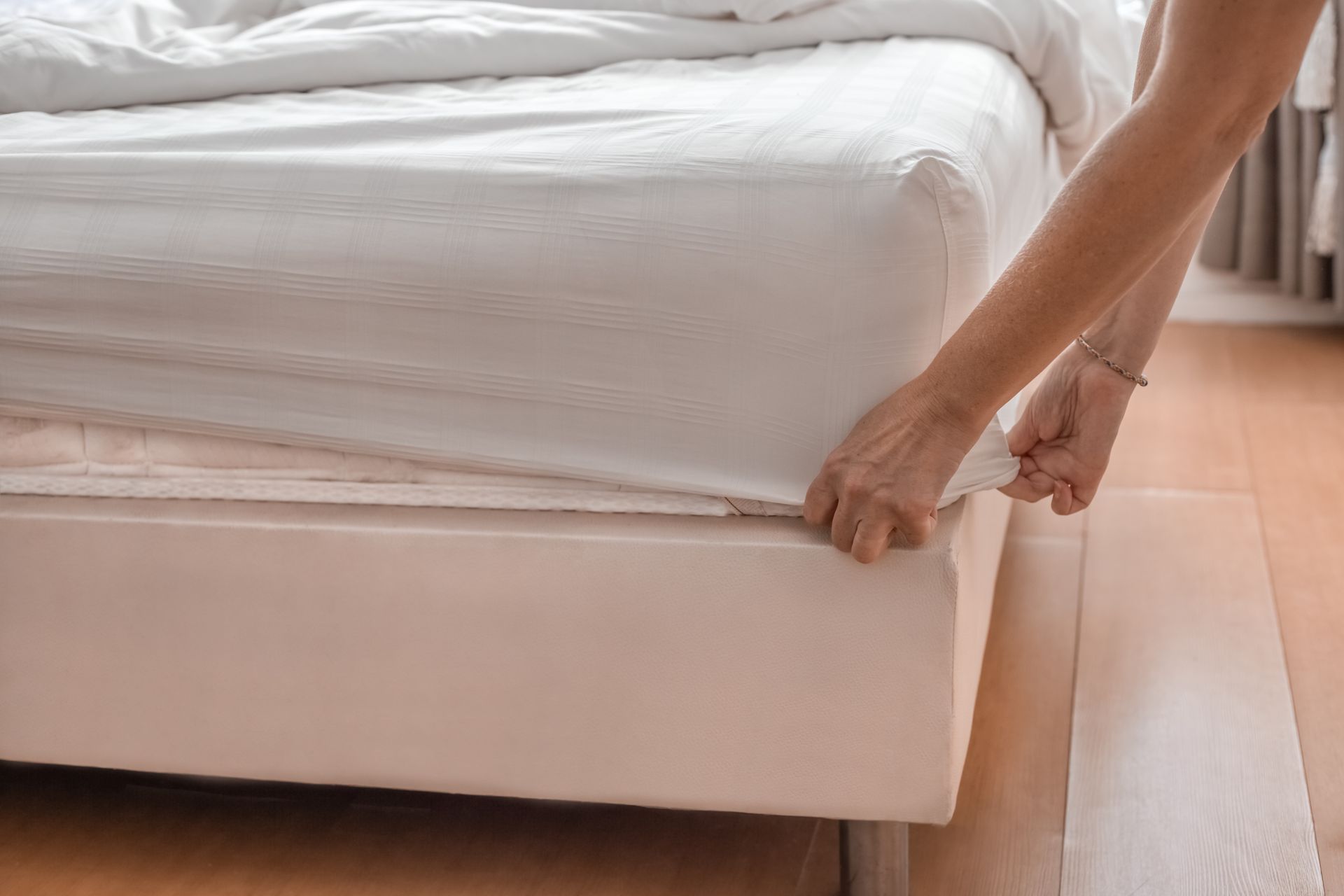 Person tucking a white fitted sheet onto a bed with a tan mattress.
