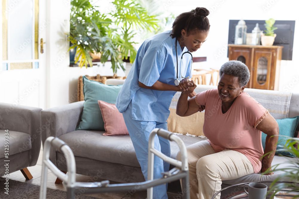Caregiver helps elderly woman rise from sofa, walker in foreground.