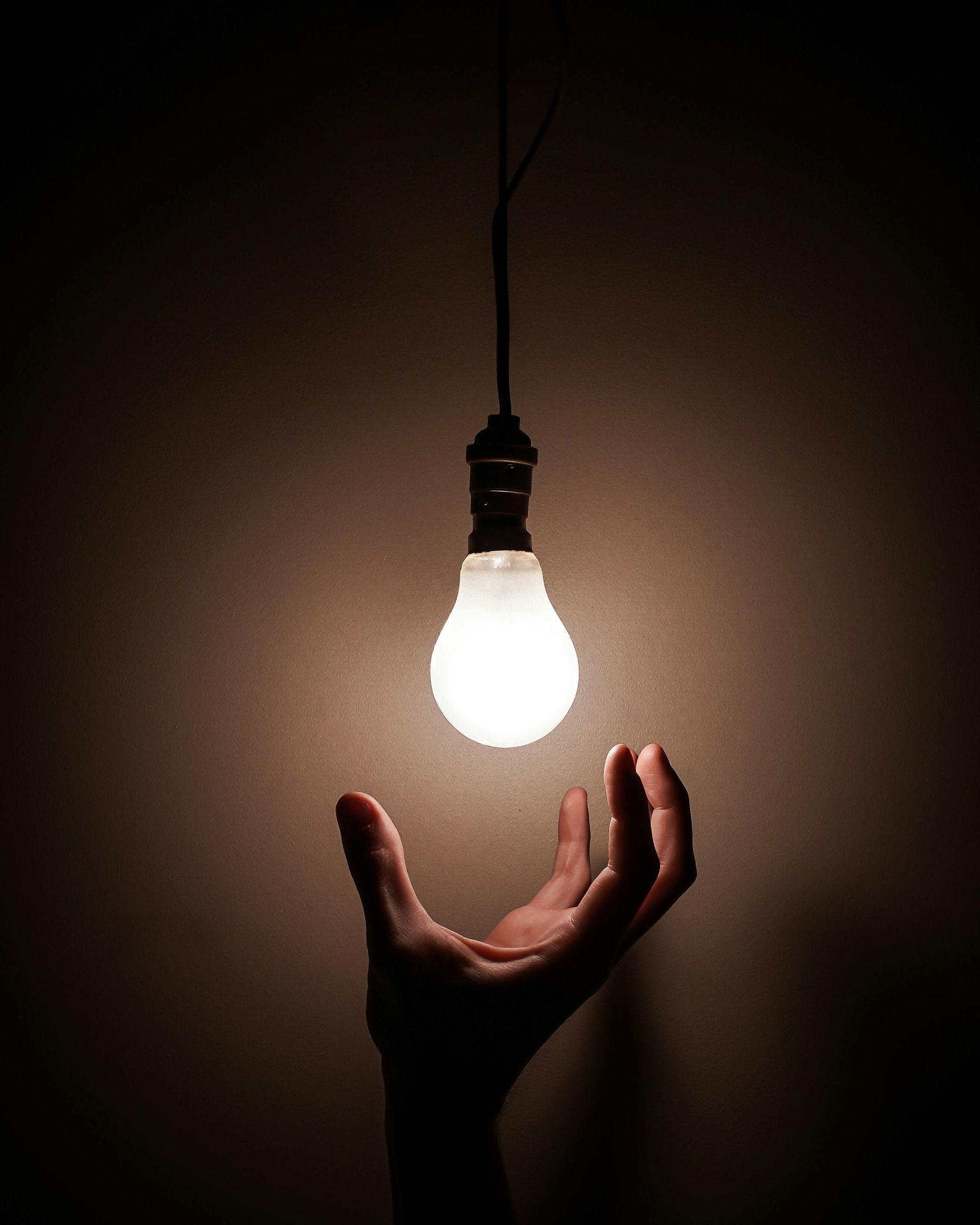 Hand reaching for a lit lightbulb suspended from a wire in a dim space.