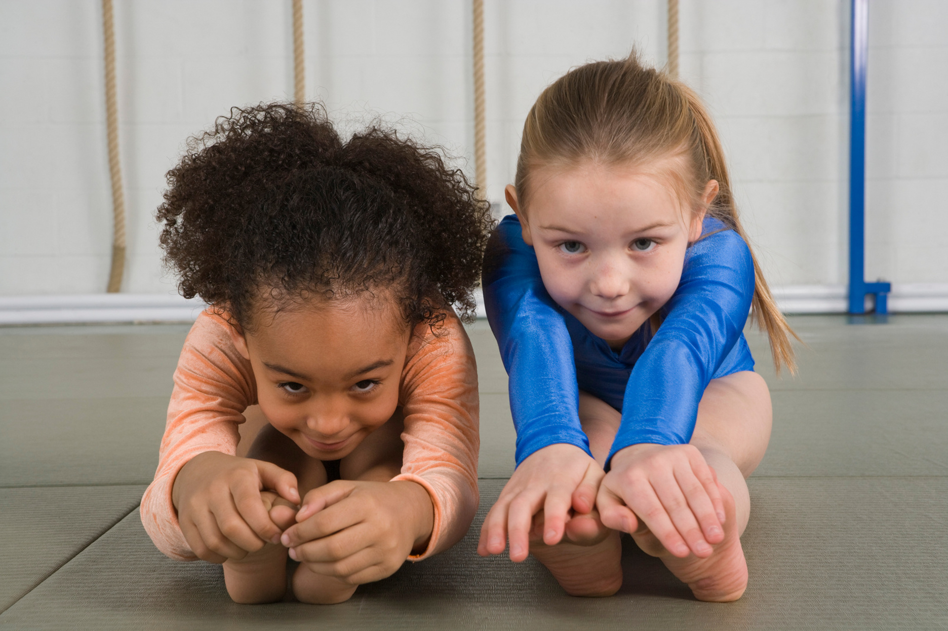 Two young girls are stretching on the floor in a gym.