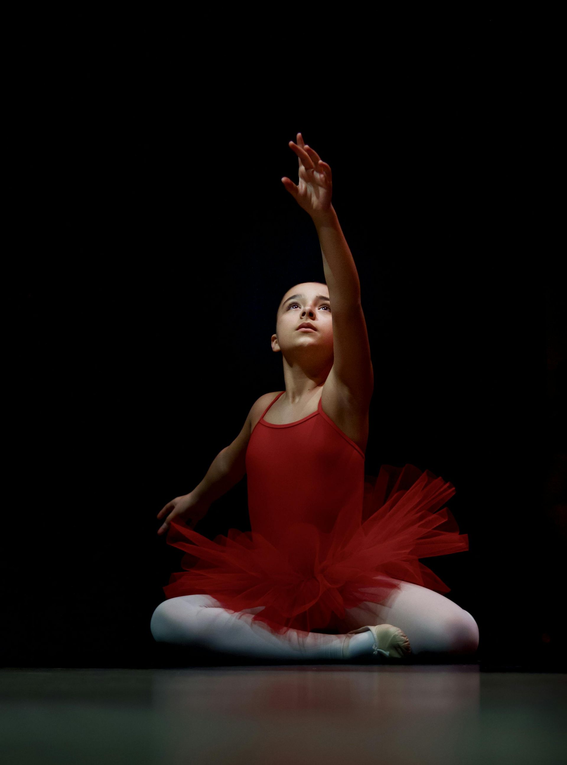 A young ballerina in a red tutu is sitting on the floor with her hands in the air.