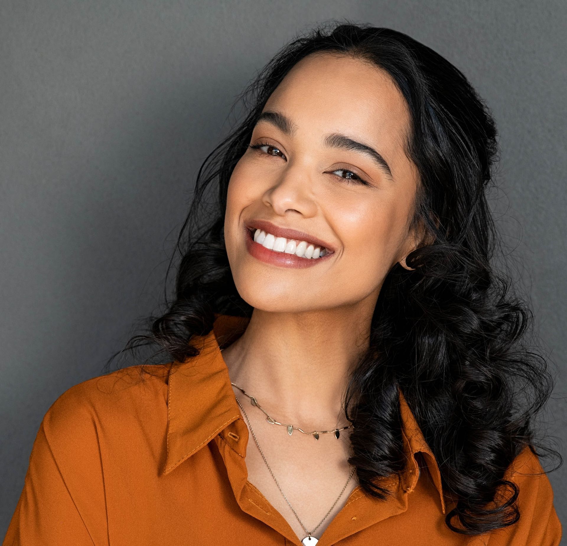 A woman wearing an orange shirt and a necklace is smiling.