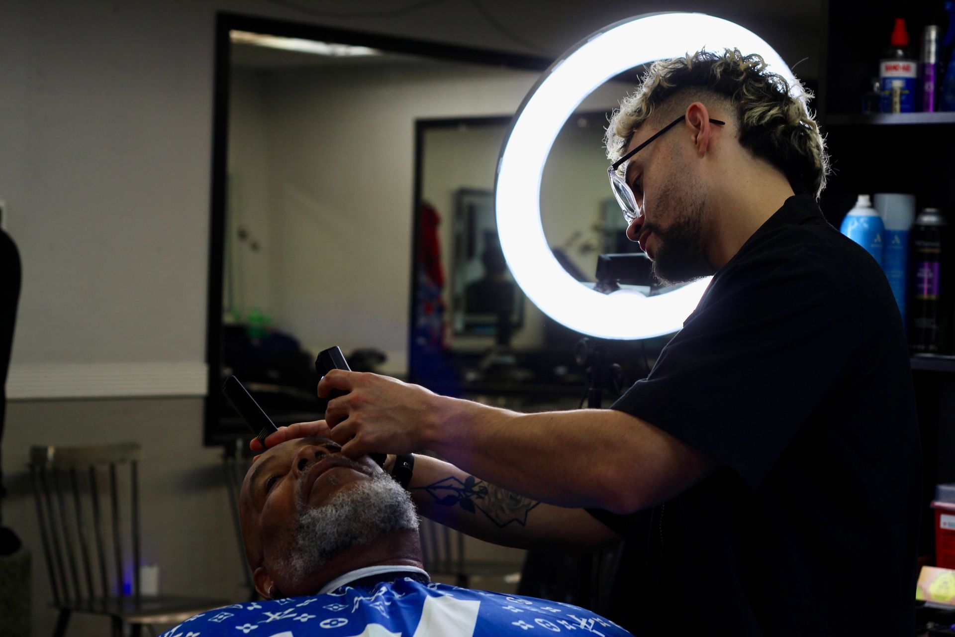 Barber shaving a client's face in a barbershop, ring light overhead.