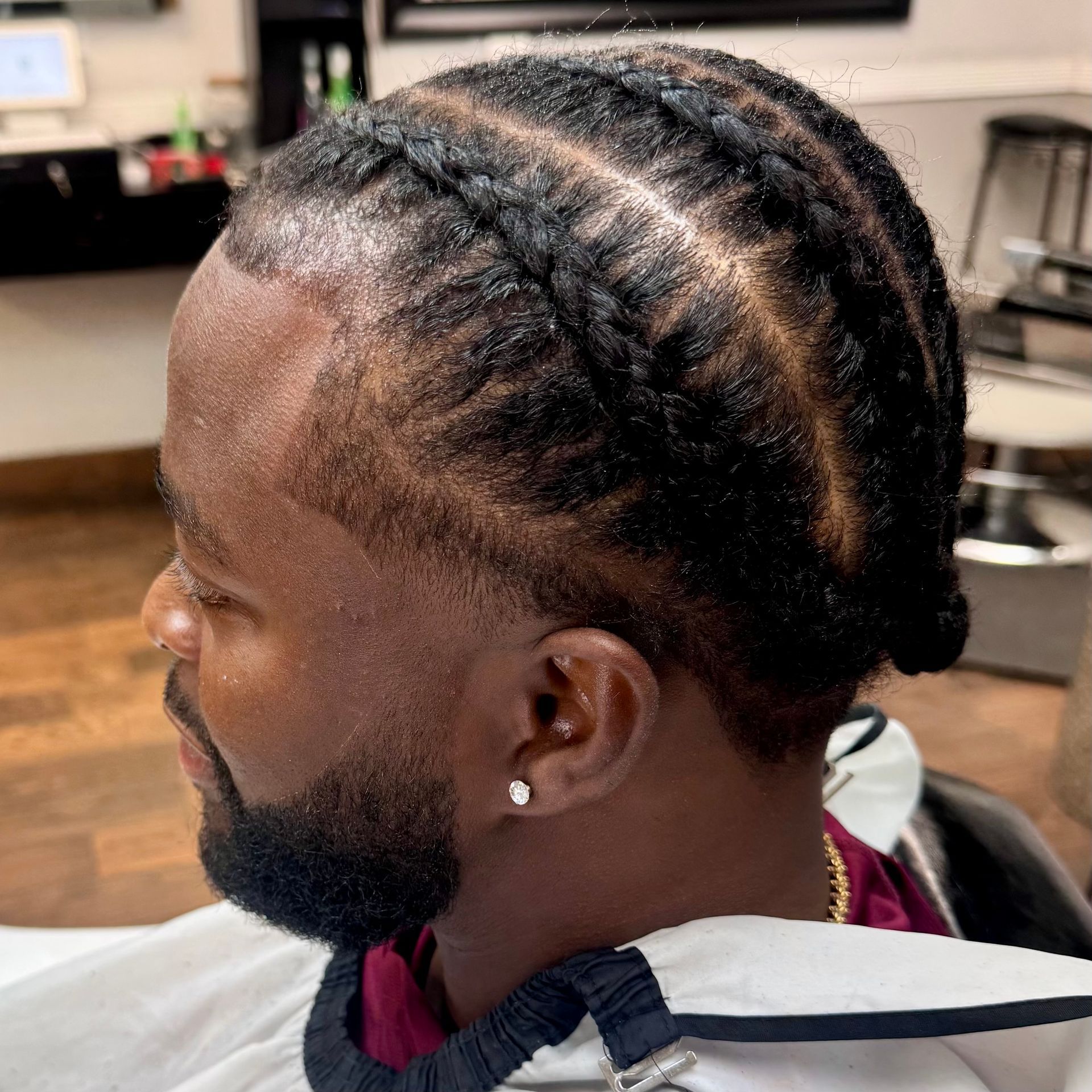 Man with cornrow braids and a trimmed beard, wearing a cape in a salon.