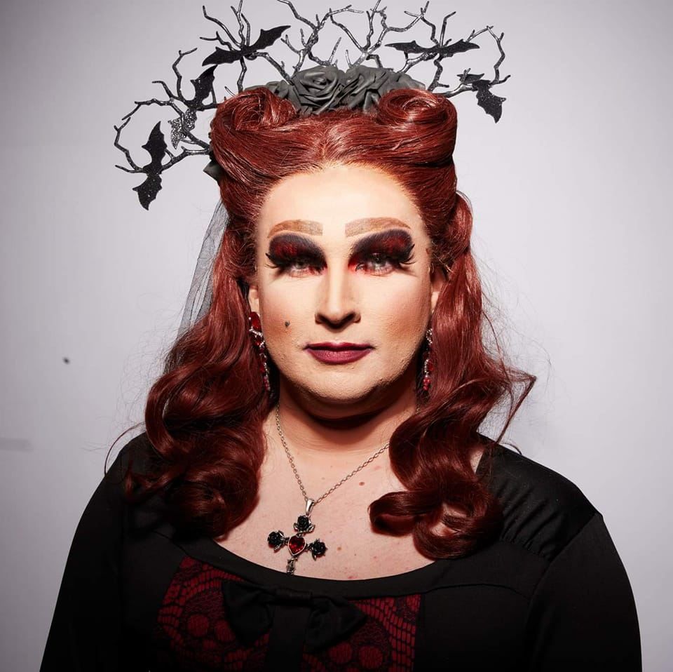 A portrait of drag queen, Evilyn Carnate (pronounced: Evil-In, Car-nate). She wears a gothic-style  make-up and outfit with the colours black and dark-red as her signature. She also has dark red hair with victory rolls to look like horns, and a headpiece with bats stuck to creepy looking branches, as though they protrude from her head.