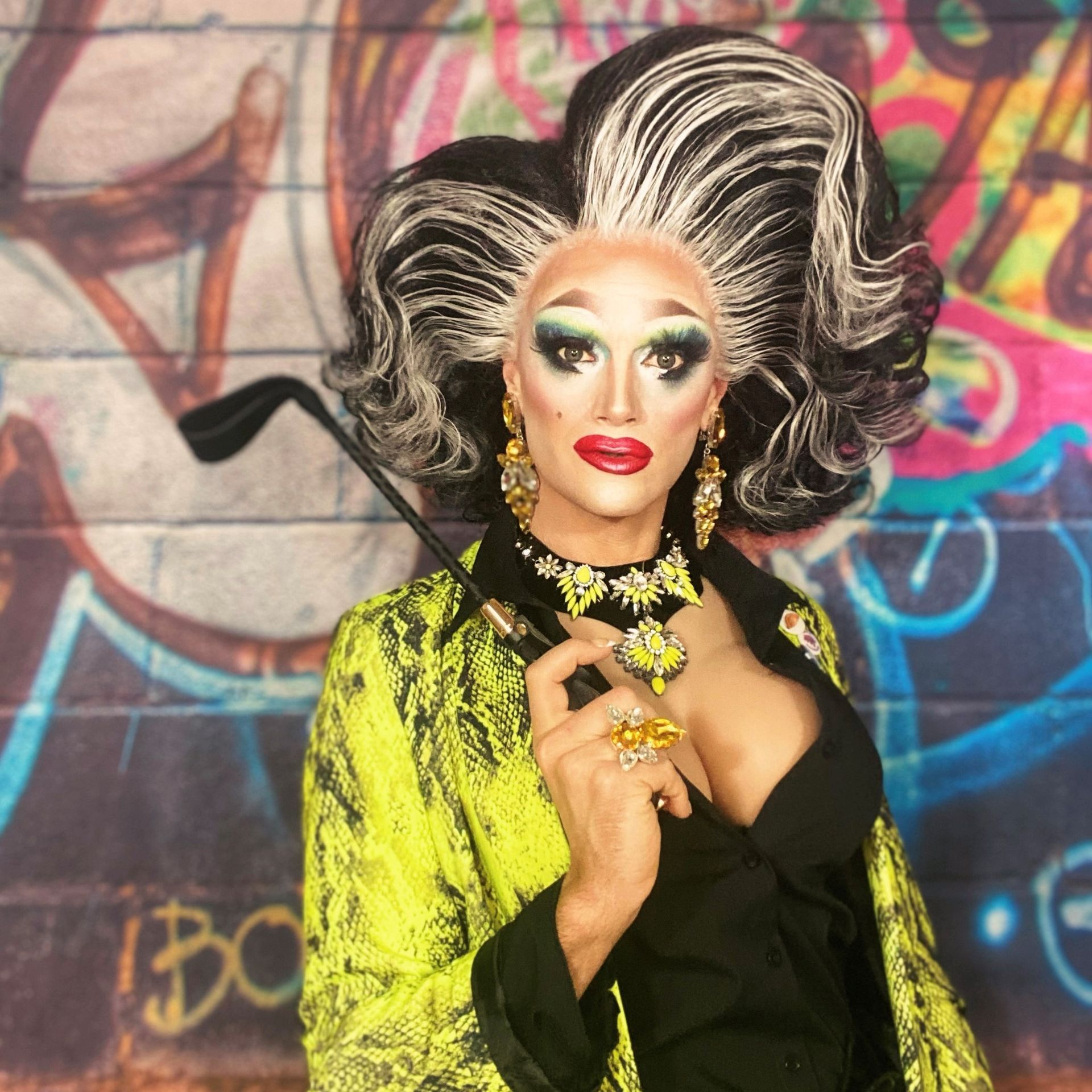 A portrait of drag queen, Felicia of Cornwall. She wears a green jacket which looks like snake skin and a low-cut black top which reveals her cleavage. Her make-up uses traditional drag queen techniques, with large blue eye shadow and overdrawn red lips. Her holds a black whip in her right hand over her shoulder. Her hair is black with white highlights running through the front, it's hair-sprayed up to accentuate it's size and is parted at the side. 