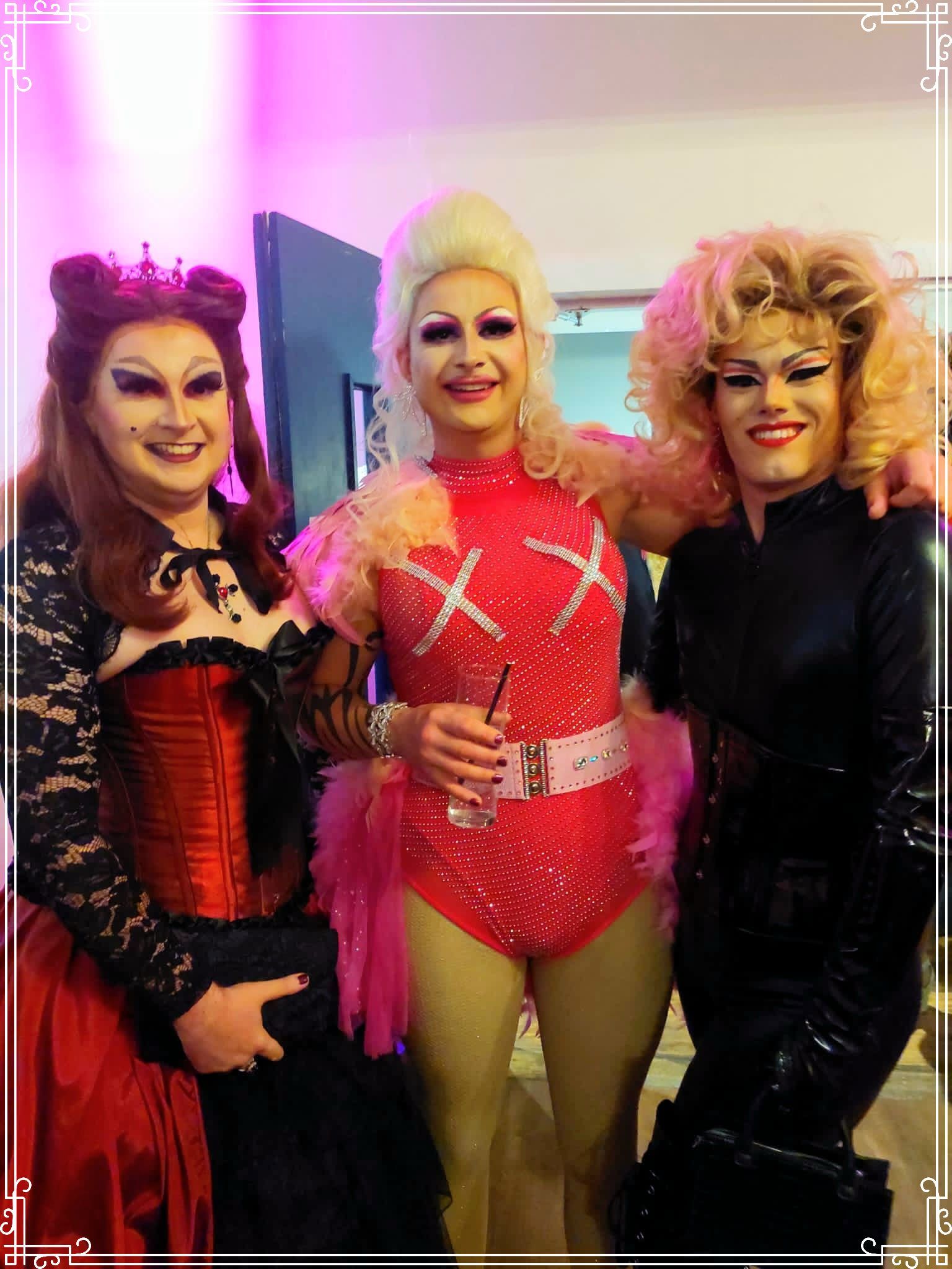 A group shot of drag queens. On the left is Evilyn Carnate (pronounced: Evil-In, Car-nate) in a gothic red and black ballgown, holding a black clutch handbag. In the middle is Rhianna Rocket, wearing a pink sequined leotard with feathers on the shoulders and hip, holding a beverage in a tall glass. On the right is Object, wearing a dominatrix catsuit with PVC gloves and corset, holding a small black handbag.