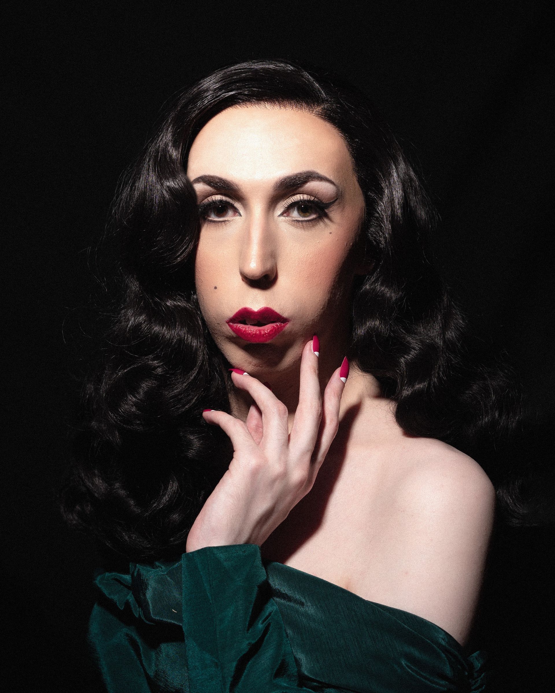 A portrait of drag queen, Roxie Moron. She wearing a vintage-style  make-up and hair in the colour black, whilst holding her hand up to her jawline to pose. Her nails are white at the base, with red tips. 
