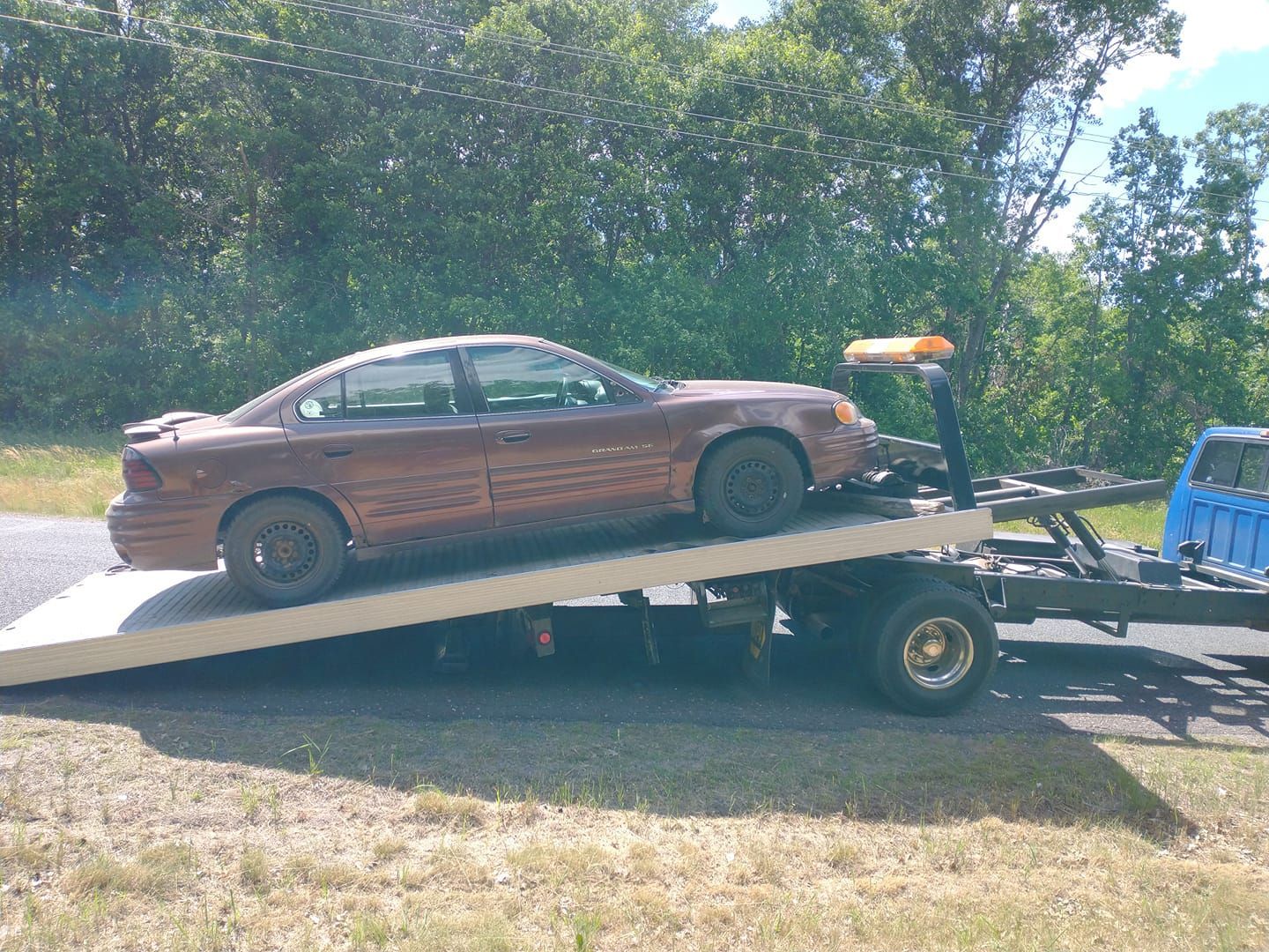Brown car being towed on a flatbed tow truck outdoors on a sunny day.