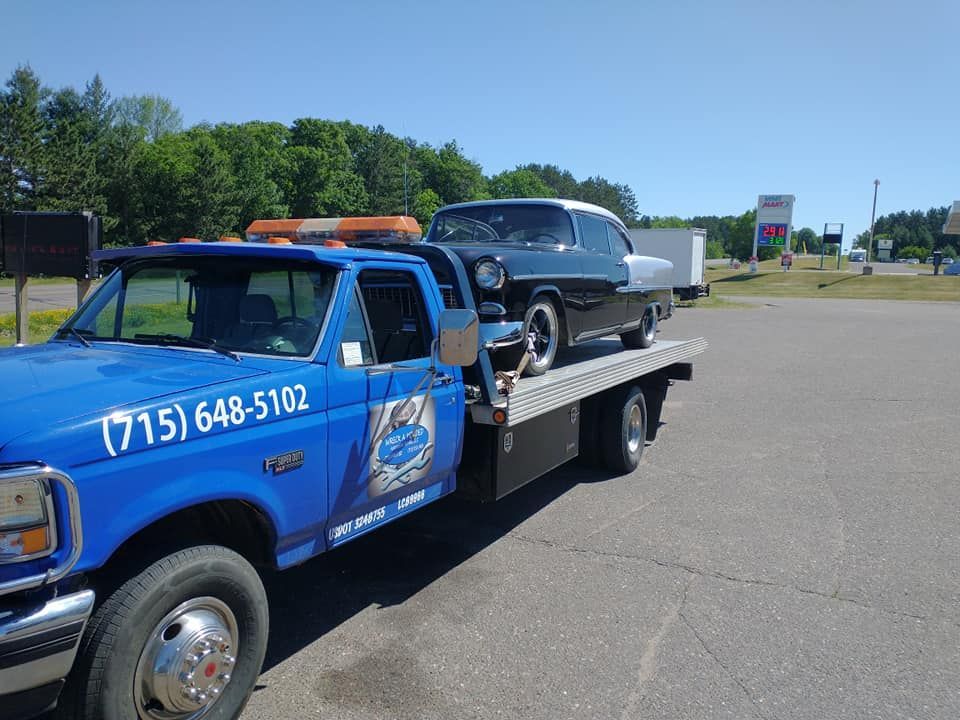 Blue tow truck carrying a classic black and white car on a sunny day.