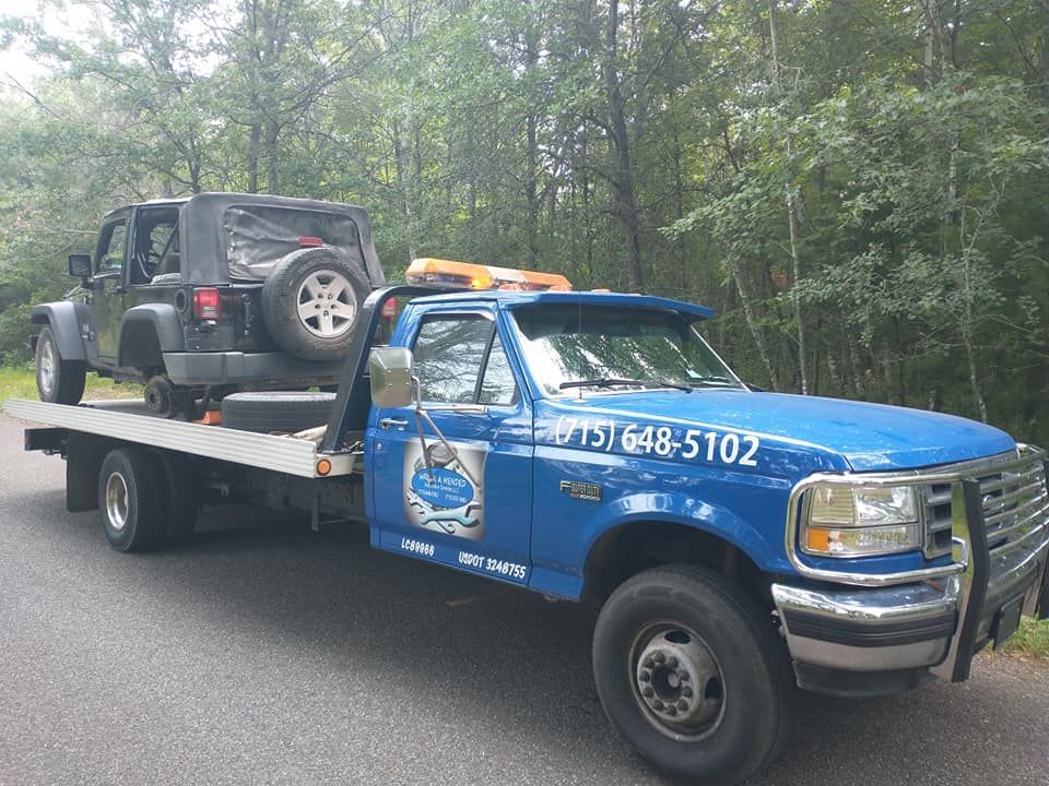Blue tow truck carrying a black Jeep on a flatbed. It is outdoors with trees in the background.