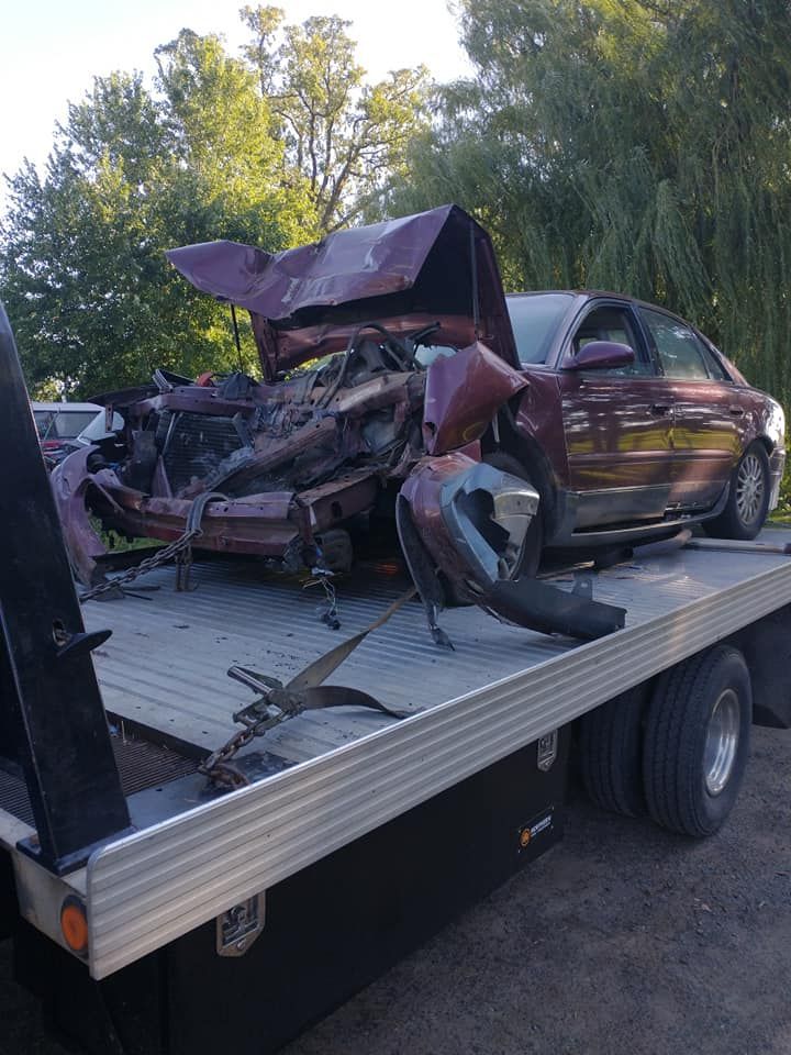A badly damaged maroon car sits on a tow truck, front end crushed, in an outdoor setting.