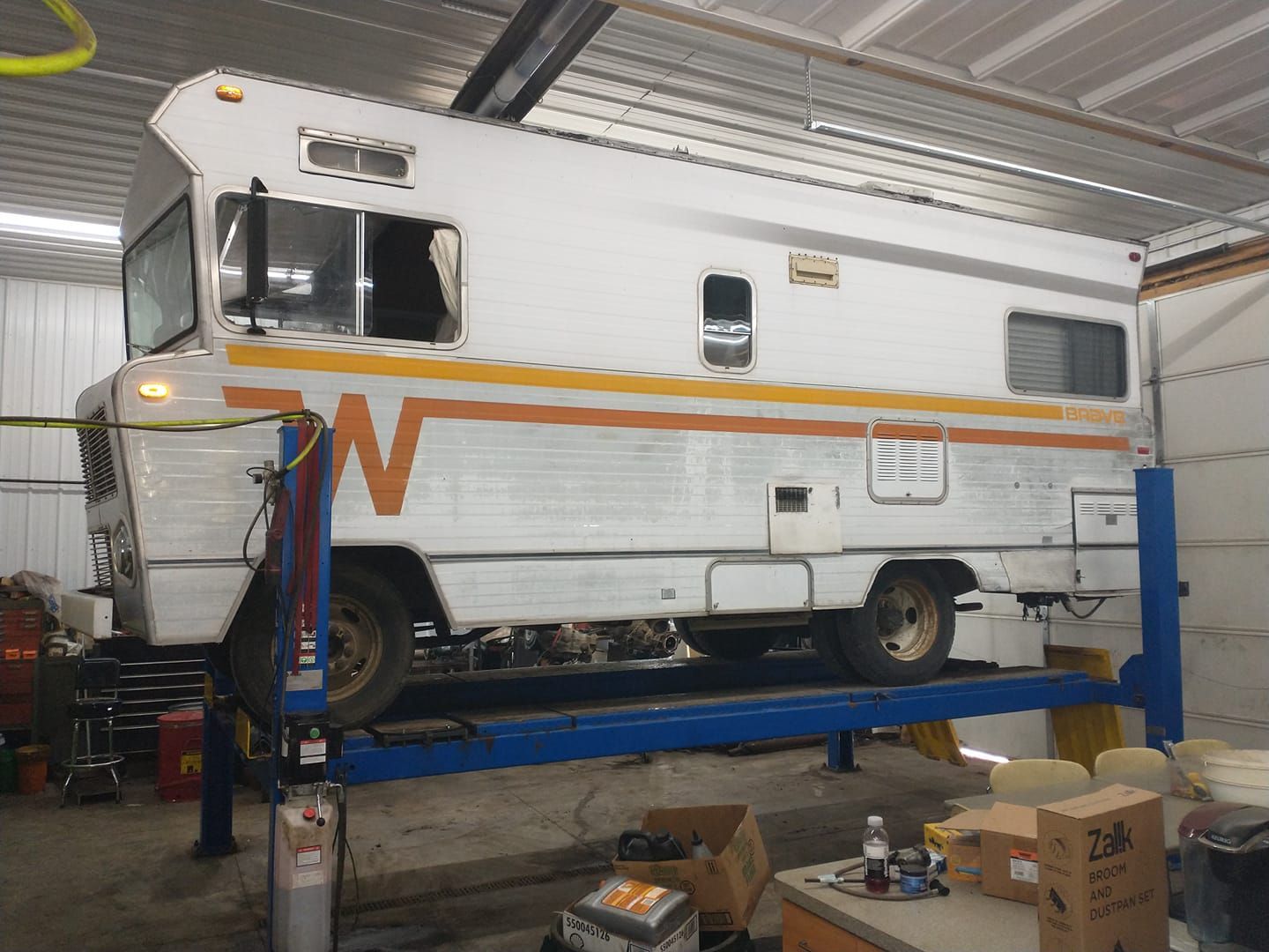 White RV on a blue lift inside a garage, with orange and yellow side stripe.