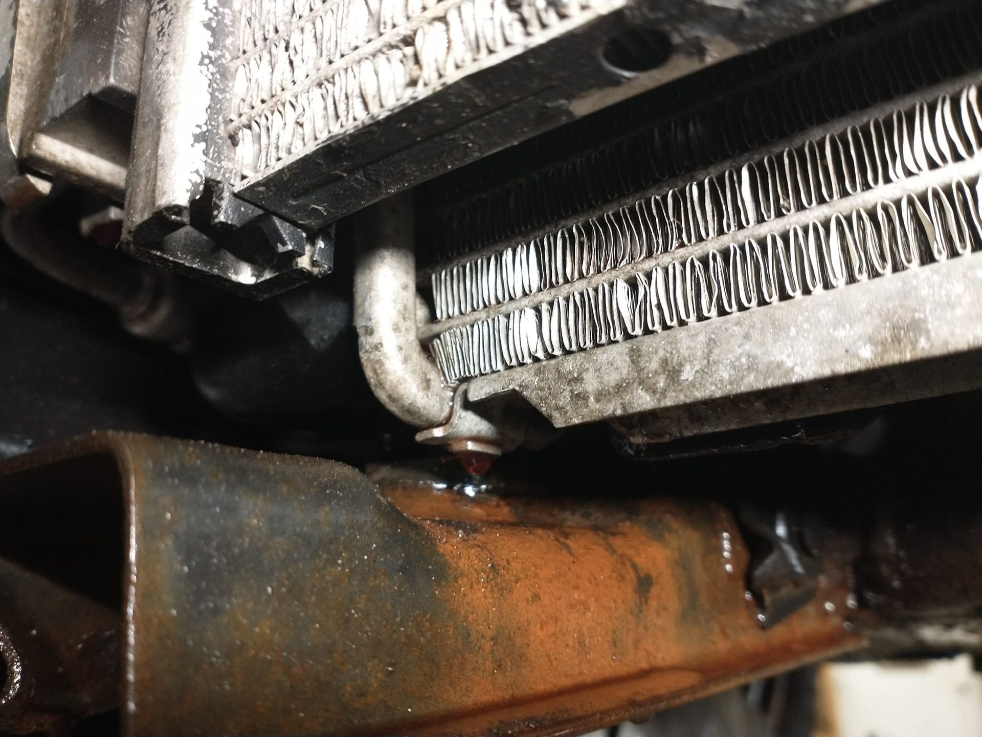 Close-up of a damaged car radiator with a dripping fluid. Rust is visible on a nearby metal frame.