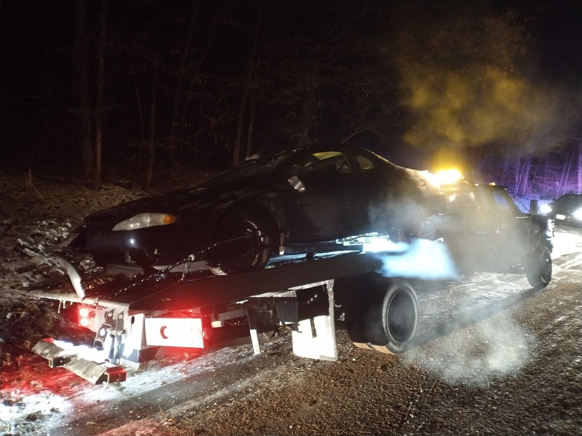 Black car on a flatbed tow truck at night, damaged with lights on; forest backdrop.