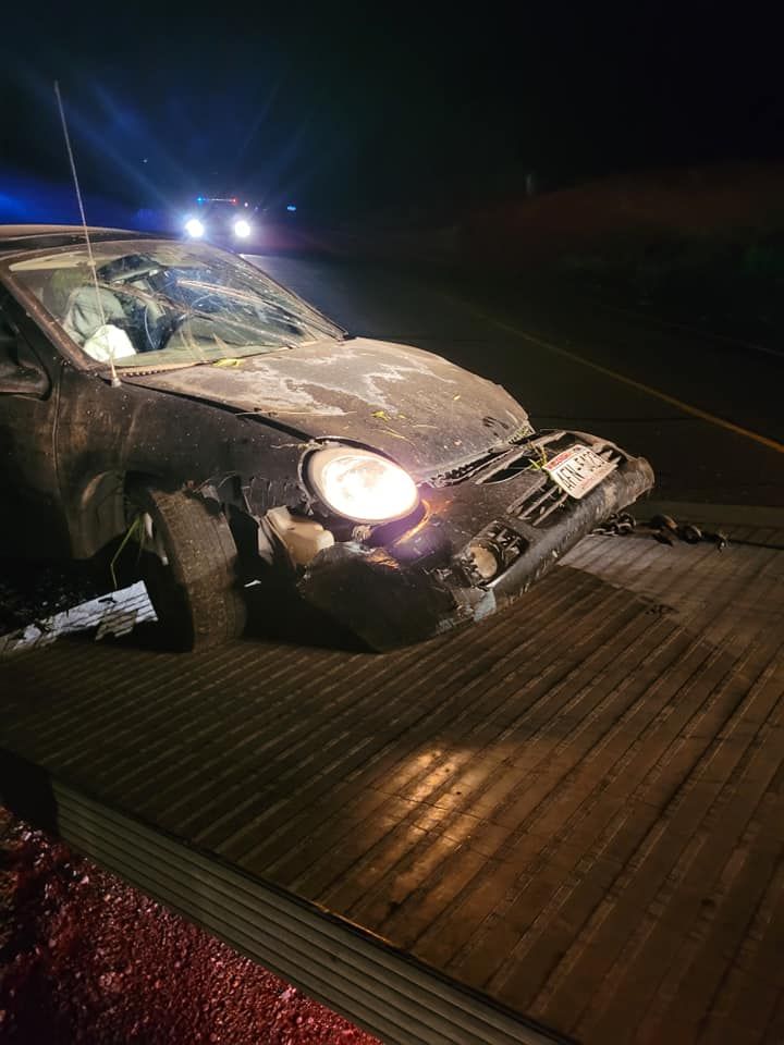 Damaged car on a tow truck at night, headlight on, front end crushed.