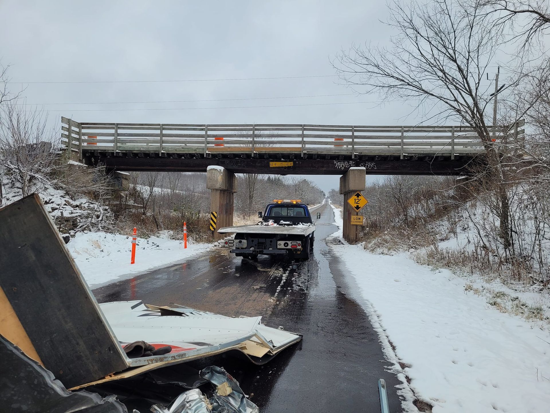 Flatbed truck under a bridge with debris on the road. Snow covers the ground, grey sky.