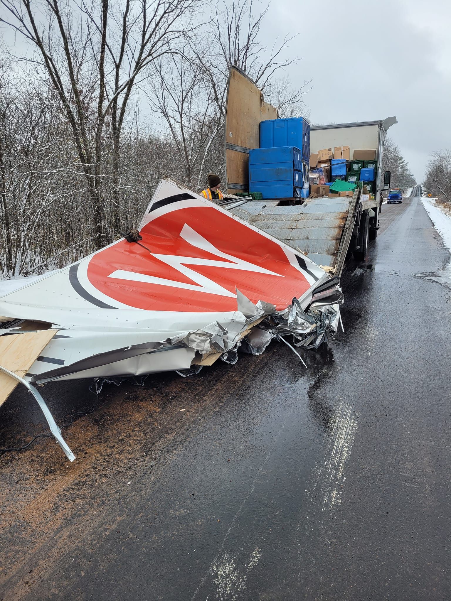 Damaged aircraft wing on a truck bed, roadside. White with red design, gray asphalt, snow-covered trees.