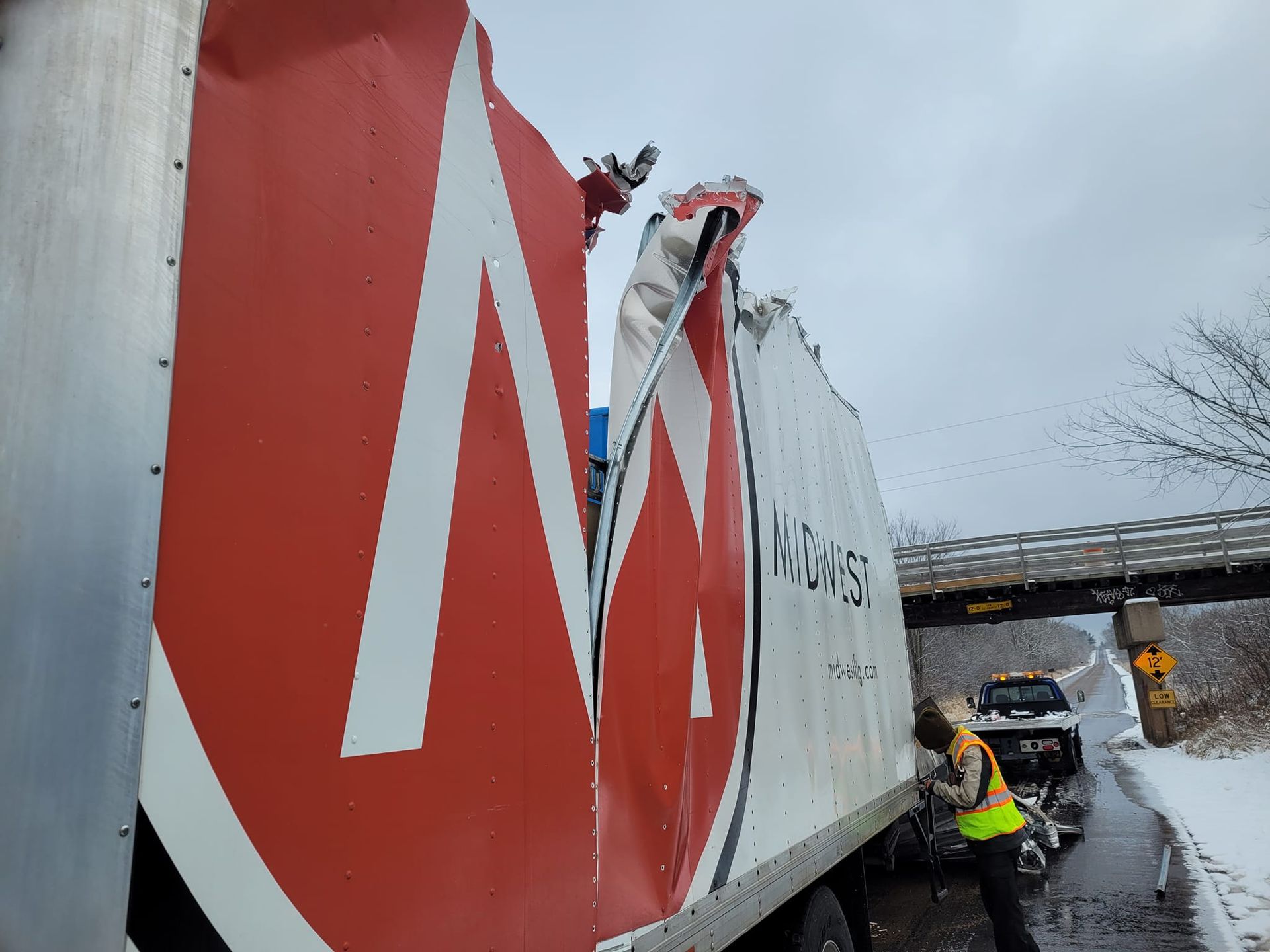Damaged semi-trailer after hitting a bridge. Red and white branding is visible. A person in a vest stands near the trailer. Snowy roadside.