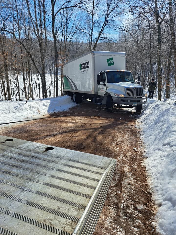 Moving truck stuck on a muddy driveway in a snowy wooded area. Two people watch.