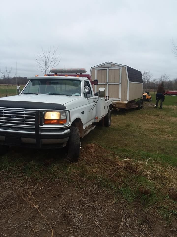 White pickup truck towing a shed on a trailer across a grassy field under an overcast sky.