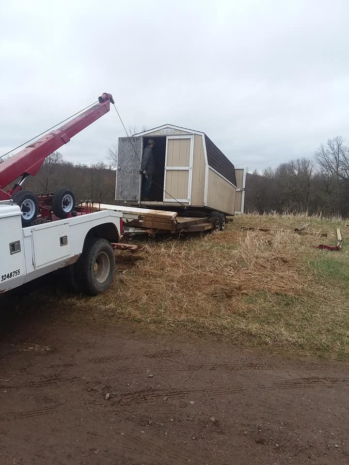 A tow truck lifting a tan and brown shed onto a flatbed trailer on a grassy field.