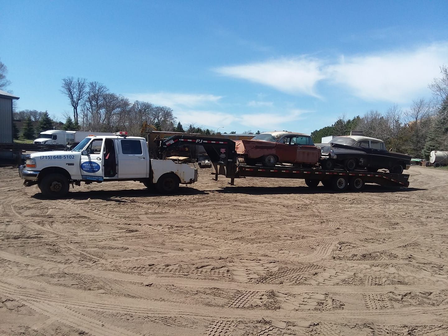 White tow truck hauling a trailer with two classic cars on a dirt lot under a blue sky.