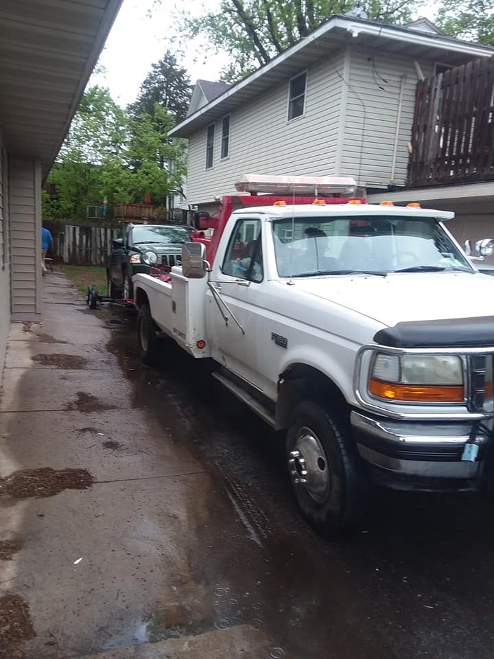 A white tow truck is towing a dark-colored car down a wet alley. A two-story building is in the background.