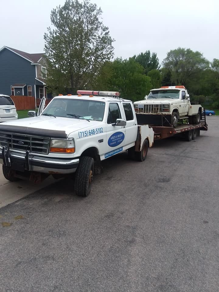 White tow truck pulling a trailer with an older white pickup truck on it; parked on a residential street.