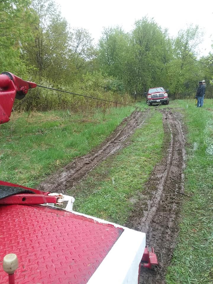 A tow truck pulling a red pickup truck stuck in muddy tracks through a grassy field. A person stands nearby.