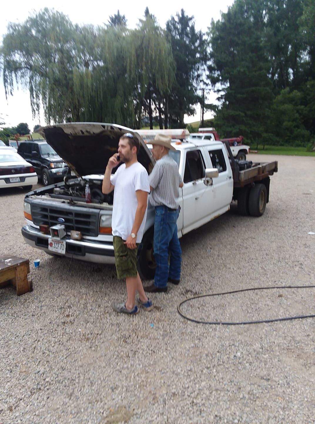 Two men beside a white pickup truck with hood up. One talks on phone, the other looks at engine.