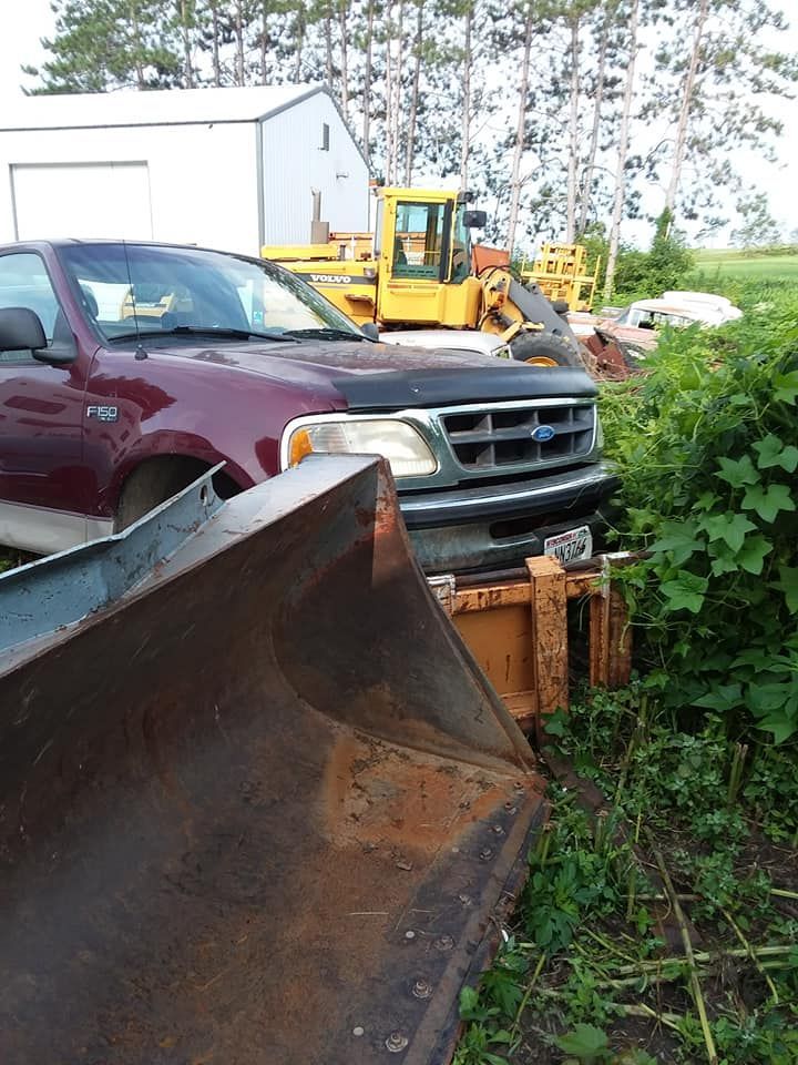 Red pickup truck with a large, rusted bucket in the foreground, with a yellow loader and building in the background.