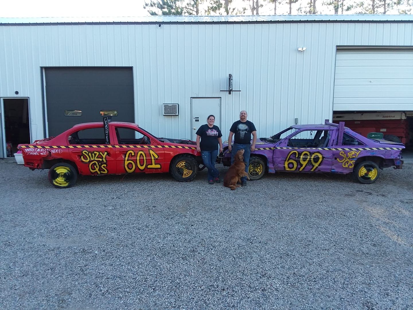Two people and dog pose beside demolition derby cars, red #601 and purple #699, outside a white building.