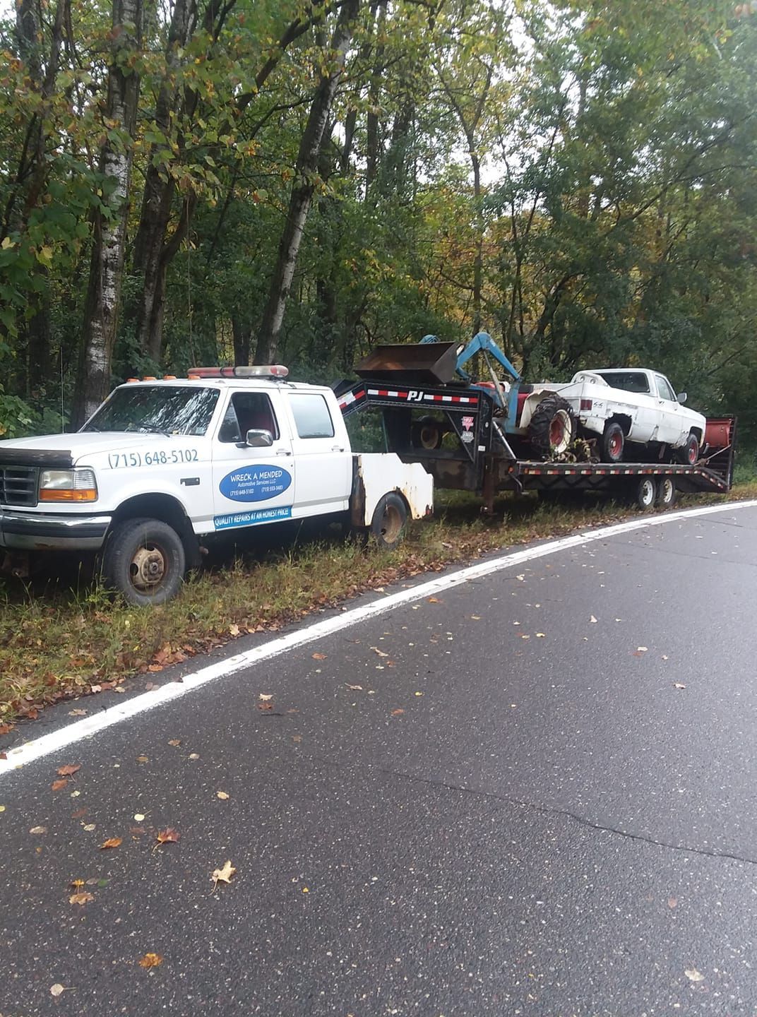 White tow truck with trailer carrying a small excavator and a white pickup truck on the side of a road.