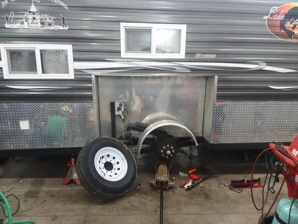 A trailer with a removed tire in a repair setting. Wheel is off, axle exposed, and a jack supports the trailer.