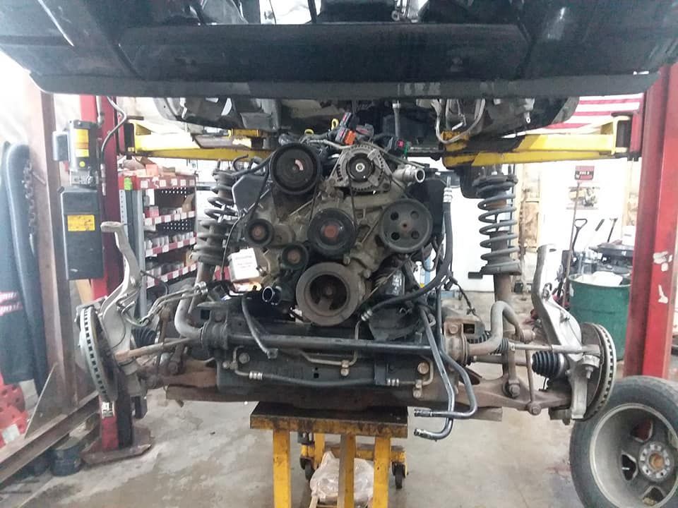 Vehicle engine and front suspension assembly lowered on a lift in a repair shop.