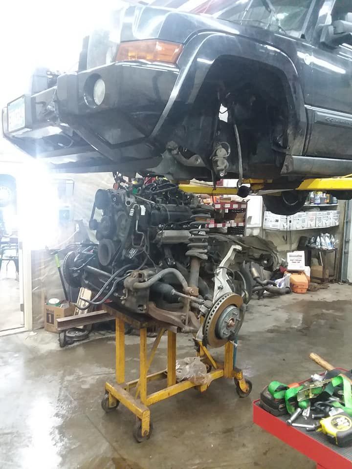 A black Jeep Commander being repaired; engine removed and on a yellow cart, in a garage.