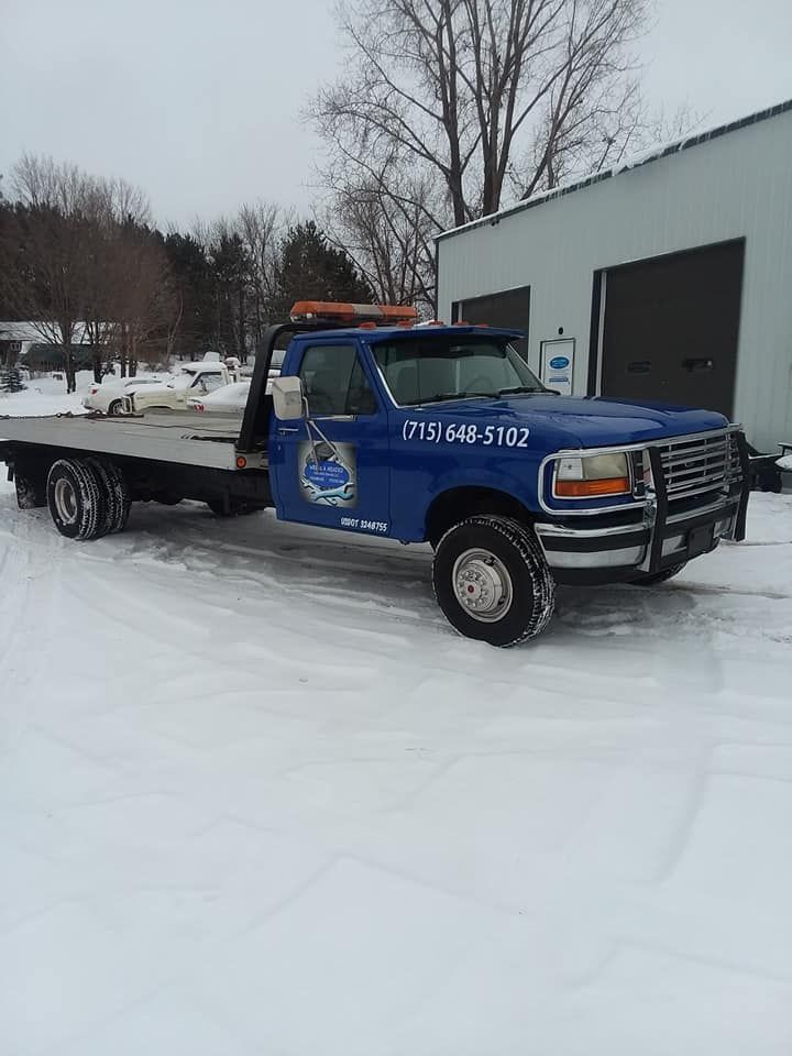 Blue tow truck parked in snow. Building and trees in the background.