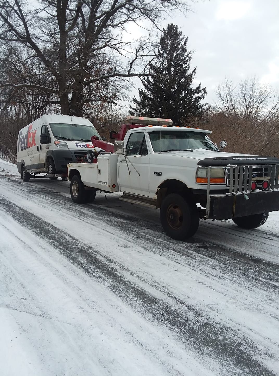 White tow truck pulling a FedEx van on a snowy road. Trees and a dark sky are in the background.