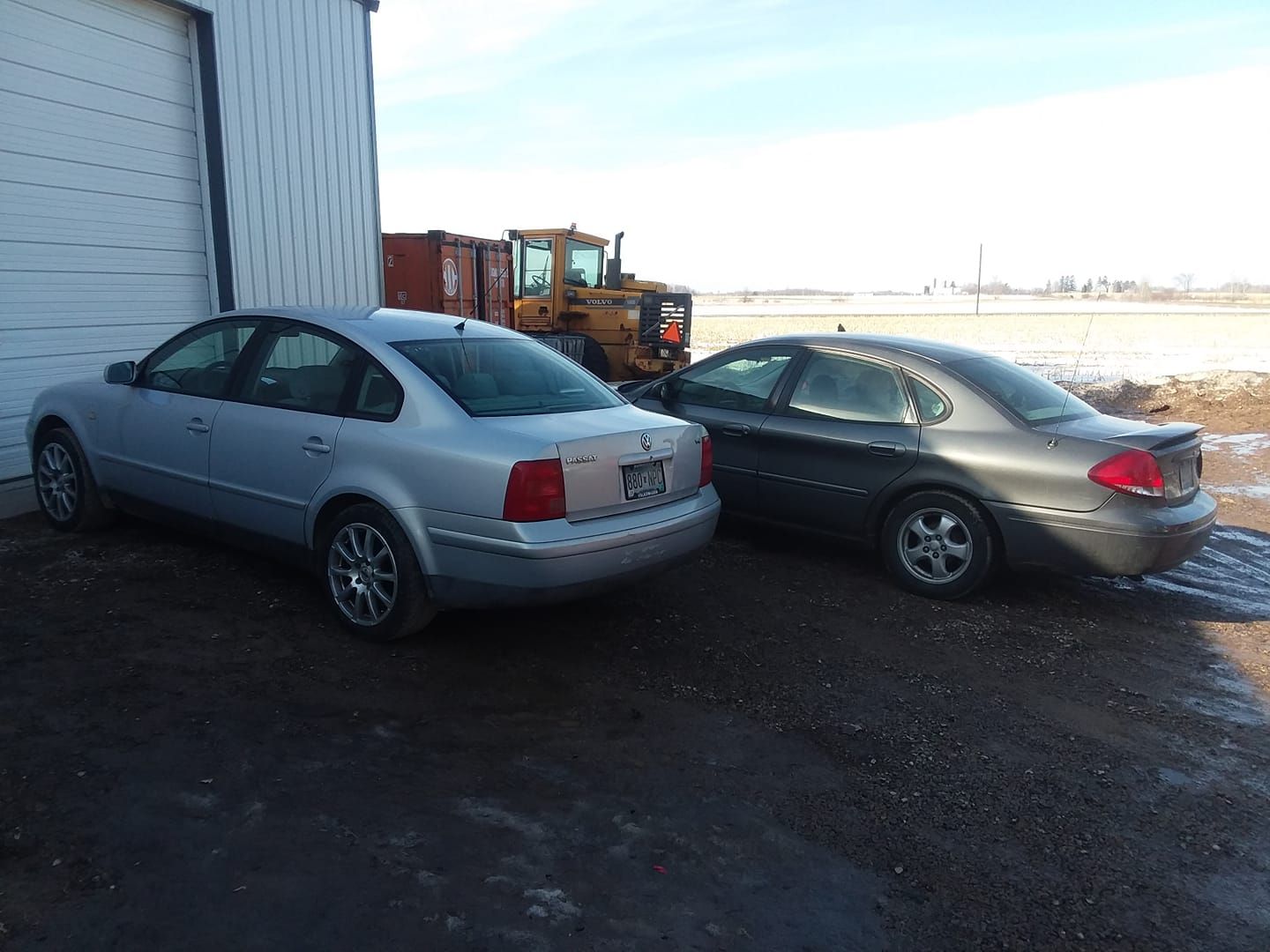 Two sedans parked next to a white building; a tractor and field are visible in the background.