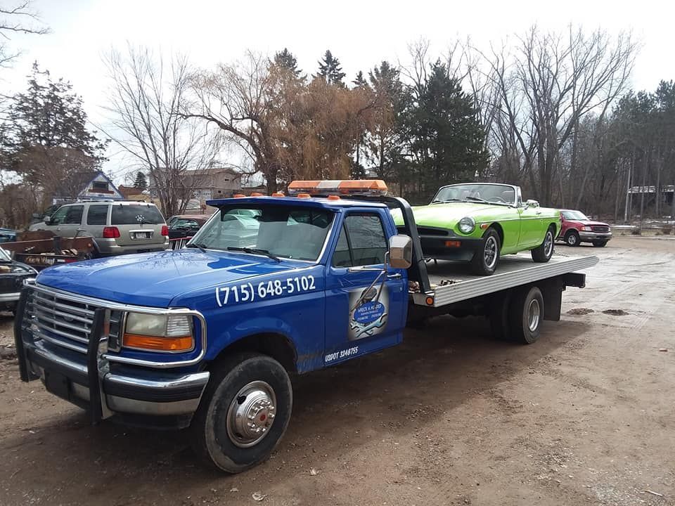 Blue tow truck with a green convertible on its flatbed in a yard.