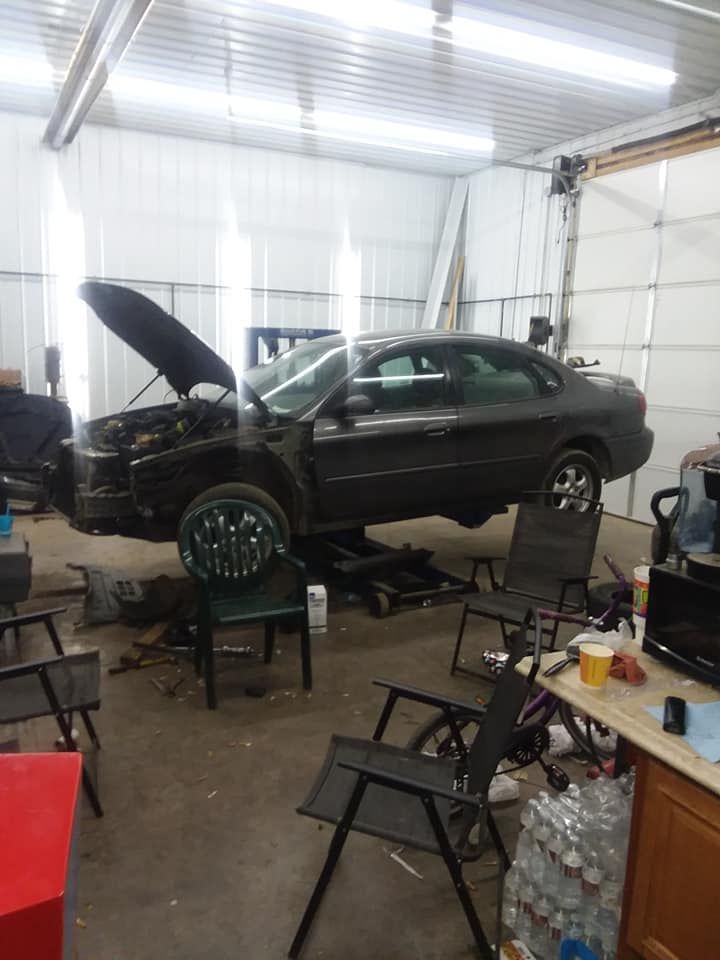 A car with its hood up is in a cluttered garage, raised on a lift.