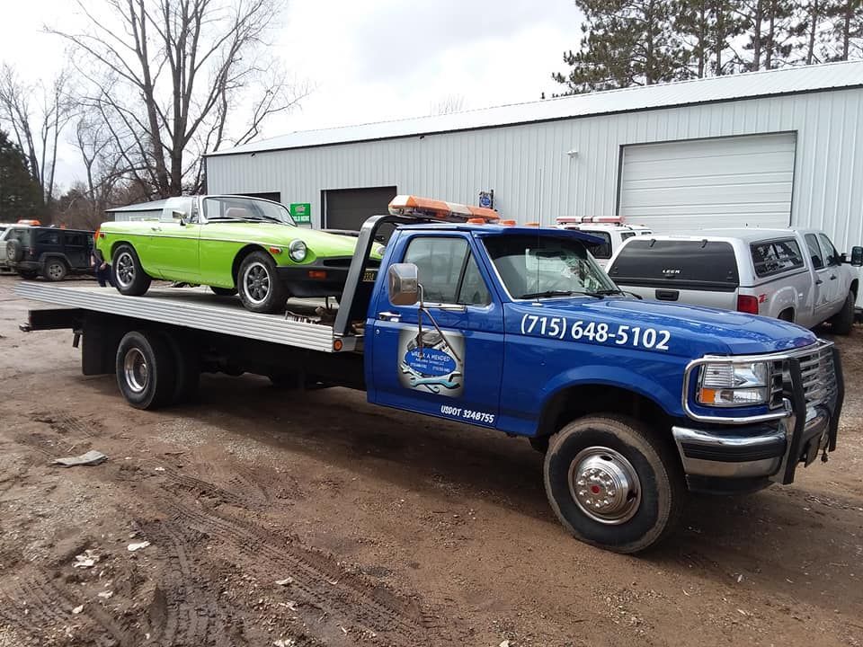 Blue tow truck transporting a green classic car at a garage. Truck has phone number (713) 648-5102.