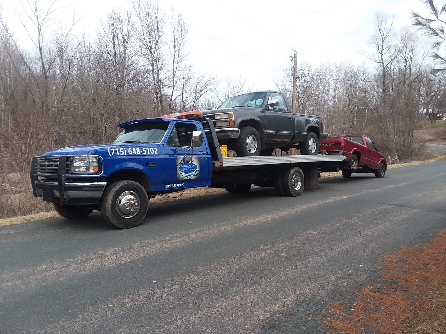 Blue tow truck carrying two damaged vehicles on a road with leafless trees.