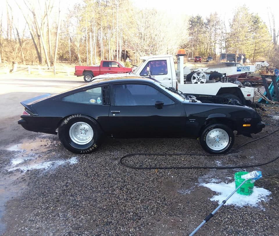 Black classic sports car parked on gravel, with a red pickup and white tow truck in background.