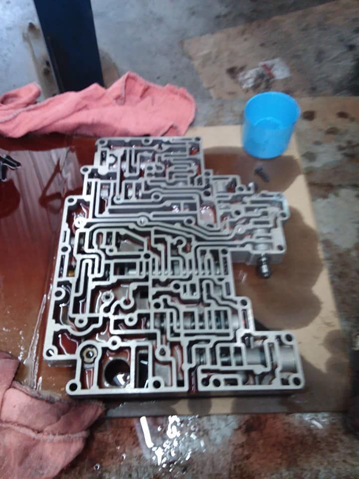 Valve body for an automatic transmission, intricate maze of channels in metal, lying on a work surface, surrounded by oil and tools.