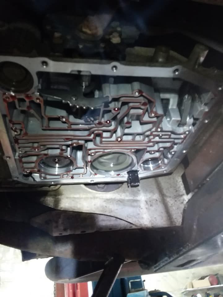 Transmission valve body inside a vehicle, viewed from below. Shows intricate channels and components with reddish fluid.