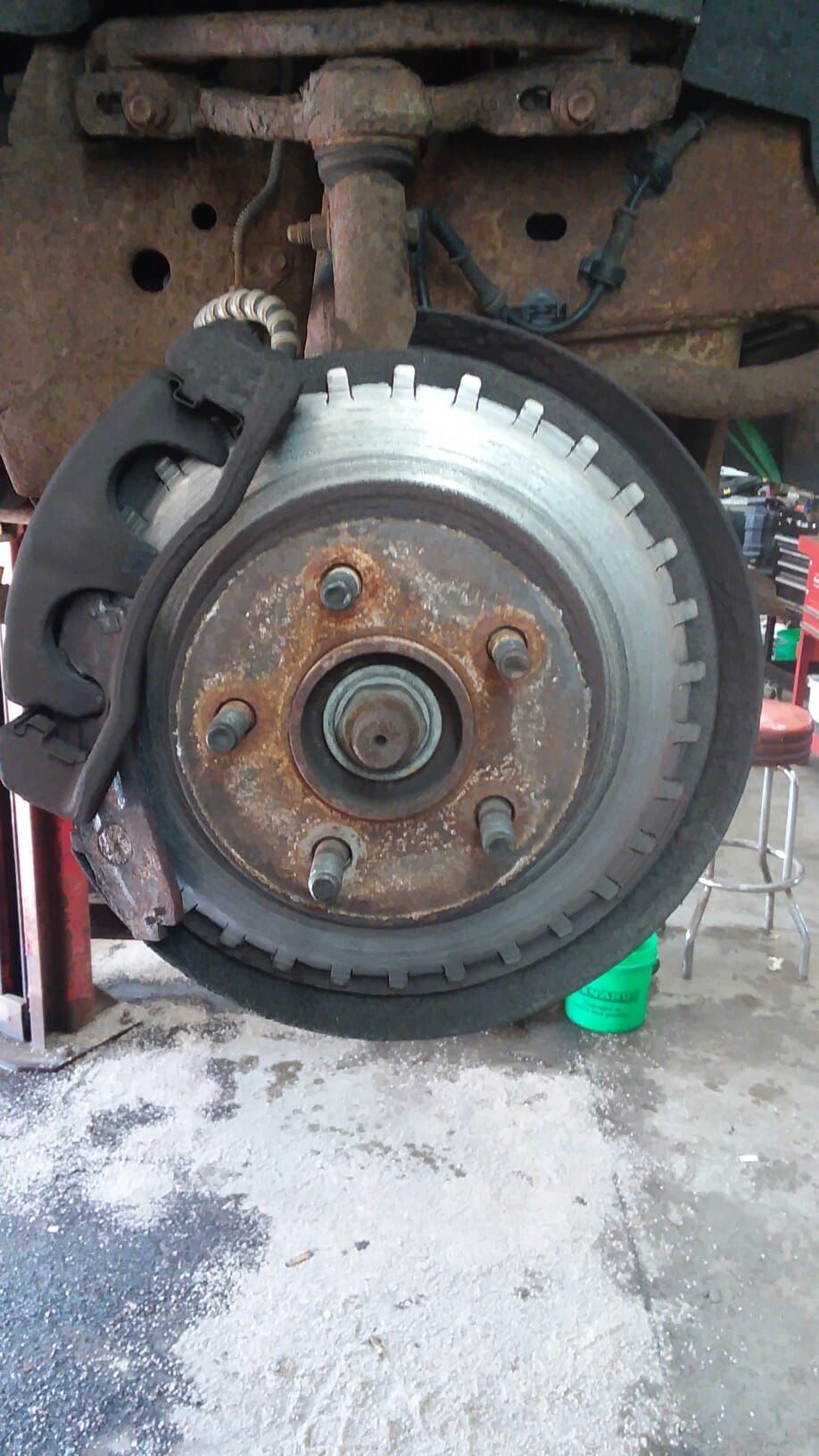 Rusty car brake rotor with caliper and mounting hardware, outdoors.