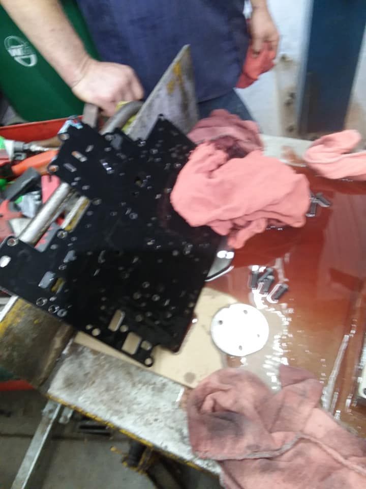 Transmission valve body on a work table, covered in oil, with red rags and tools nearby.
